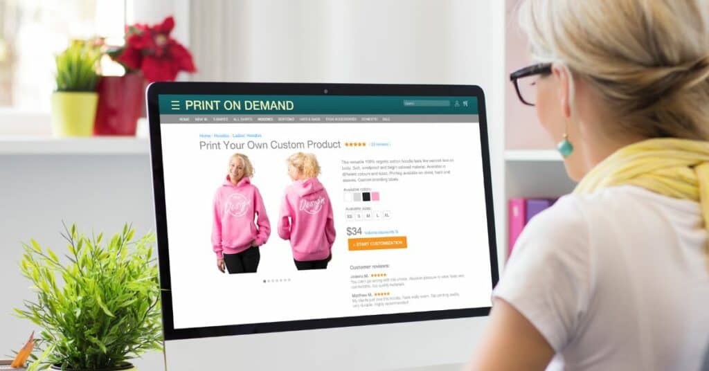 A woman examines a computer screen displaying a pink hoodie print-on-demand design.