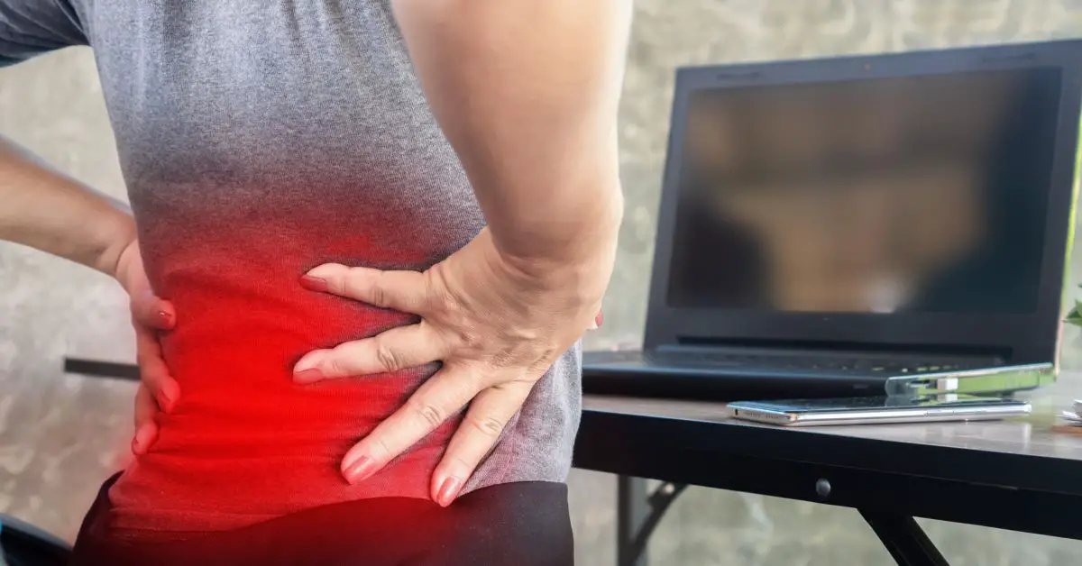 lower back pain after sitting too long - A lady sitting in front of a laptop with both hands holding her lower back