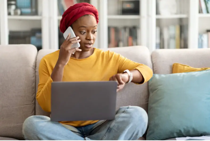 A woman effectively managing time at work while sitting on a couch with a laptop and cell phone.